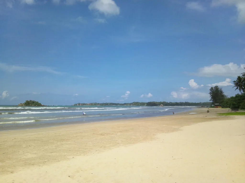 Wide sandy Weligama Beach with low breaking waves and the bay beneath a blue sky, Sri Lanka